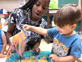 A teacher smiles while a young male student holds up his painting.